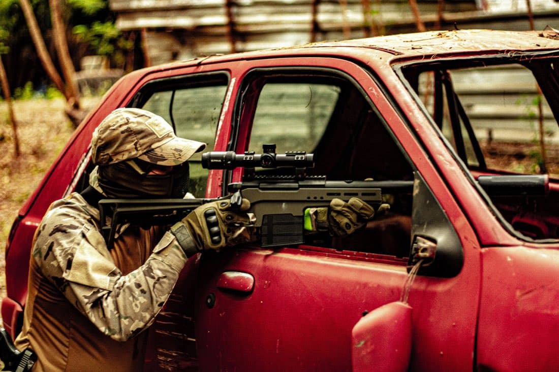 Operador mirando com rifle de airsoft apoiado em um carro abandonado no campo
